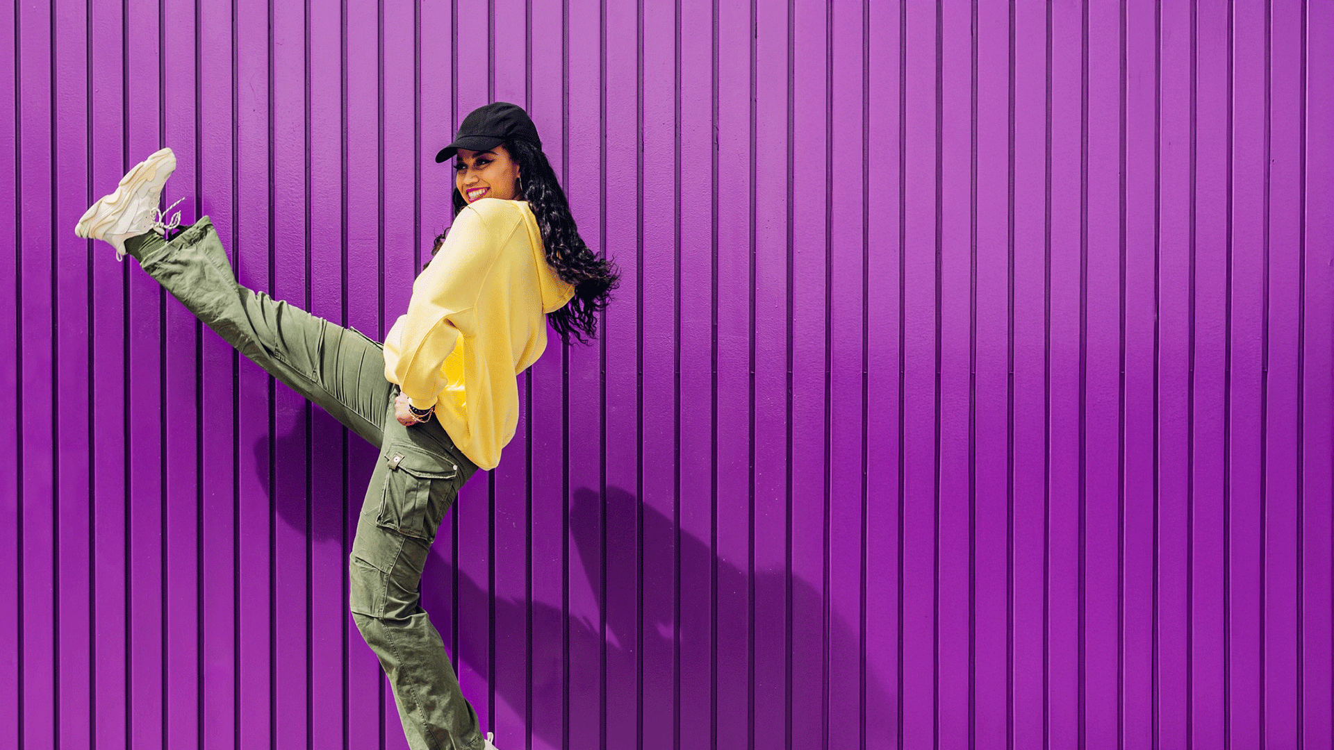 Young woman in a yellow top kicking up her heels against a bright purple wall